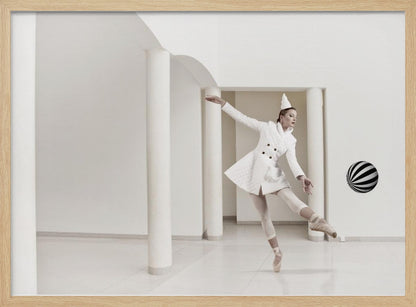 A ballerina dressed in a white quilted coat and a pointed hat dances en pointe in a minimalist white hall with large columns. She gracefully extends her arms, looking towards a black and white striped ball that appears to be floating in mid-air. The scene is presented within a silver picture frame. Wall Art