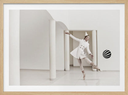 A ballerina dressed in a white quilted coat and a pointed hat dances en pointe in a minimalist white hall with large columns. She gracefully extends her arms, looking towards a black and white striped ball that appears to be floating in mid-air. The scene is presented within a silver picture frame. Wall Art