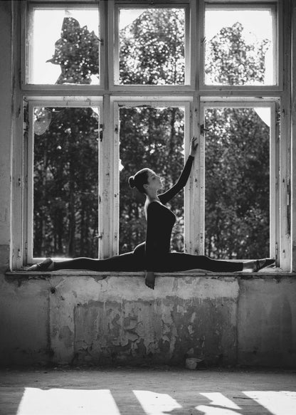 A black and white photograph of a graceful female dancer holding a perfect split on a large, dilapidated windowsill. She is silhouetted against the bright light from the broken window panes, with her arm elegantly raised. The setting is an abandoned room with peeling paint, creating a stark contrast between the dancer's elegance and the decaying environment. Wall Art