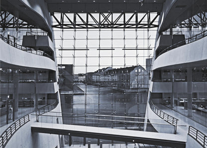 A symmetrical, black and white photograph taken from inside a modern building with curved balconies, looking out through a massive glass wall onto a canal and historic buildings on the opposite bank. A pedestrian bridge crosses the lower part of the frame. Decor
