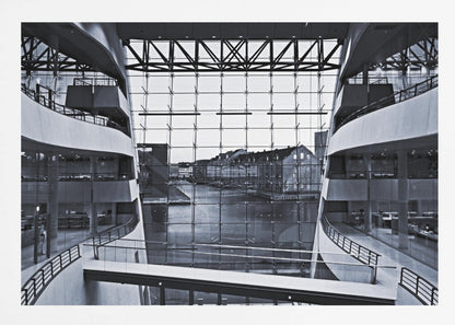 A symmetrical, black and white photograph taken from inside a modern building with curved balconies, looking out through a massive glass wall onto a canal and historic buildings on the opposite bank. A pedestrian bridge crosses the lower part of the frame. Decor