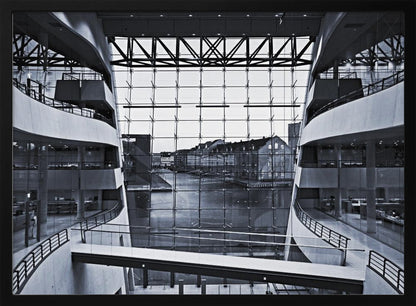 A symmetrical, black and white photograph taken from inside a modern building with curved balconies, looking out through a massive glass wall onto a canal and historic buildings on the opposite bank. A pedestrian bridge crosses the lower part of the frame. Decor