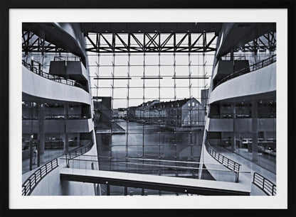 A symmetrical, black and white photograph taken from inside a modern building with curved balconies, looking out through a massive glass wall onto a canal and historic buildings on the opposite bank. A pedestrian bridge crosses the lower part of the frame. Decor