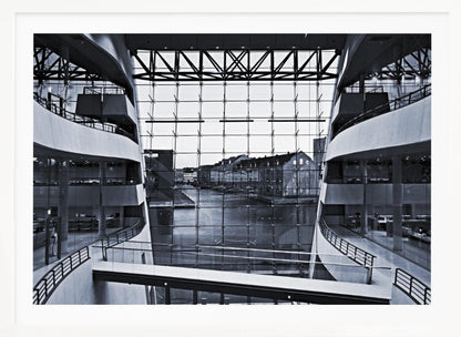 A symmetrical, black and white photograph taken from inside a modern building with curved balconies, looking out through a massive glass wall onto a canal and historic buildings on the opposite bank. A pedestrian bridge crosses the lower part of the frame. Decor