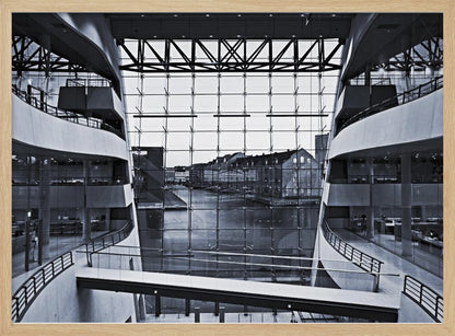 A symmetrical, black and white photograph taken from inside a modern building with curved balconies, looking out through a massive glass wall onto a canal and historic buildings on the opposite bank. A pedestrian bridge crosses the lower part of the frame. Decor