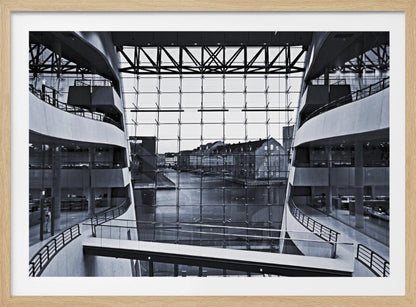 A symmetrical, black and white photograph taken from inside a modern building with curved balconies, looking out through a massive glass wall onto a canal and historic buildings on the opposite bank. A pedestrian bridge crosses the lower part of the frame. Decor