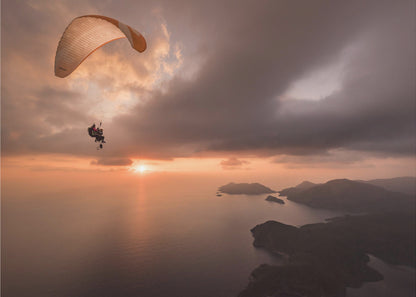 A scenic aerial view of a person paragliding over the ocean during a dramatic sunset. The orange and white parachute is in the upper left corner against a cloudy sky. The sun on the horizon casts a warm glow on the water, with silhouetted islands in the distance. Poster