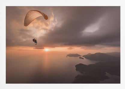 A scenic aerial view of a person paragliding over the ocean during a dramatic sunset. The orange and white parachute is in the upper left corner against a cloudy sky. The sun on the horizon casts a warm glow on the water, with silhouetted islands in the distance. Poster