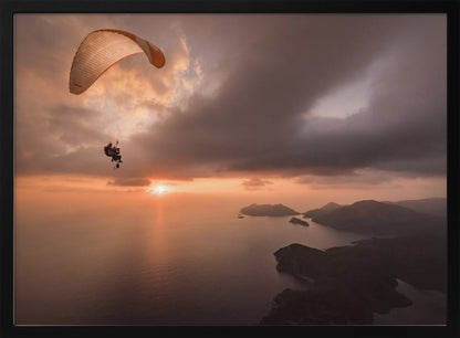 A scenic aerial view of a person paragliding over the ocean during a dramatic sunset. The orange and white parachute is in the upper left corner against a cloudy sky. The sun on the horizon casts a warm glow on the water, with silhouetted islands in the distance. Poster
