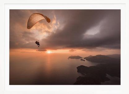 A scenic aerial view of a person paragliding over the ocean during a dramatic sunset. The orange and white parachute is in the upper left corner against a cloudy sky. The sun on the horizon casts a warm glow on the water, with silhouetted islands in the distance. Poster