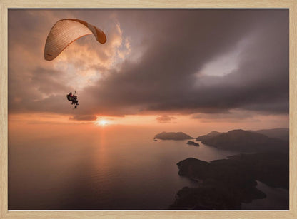A scenic aerial view of a person paragliding over the ocean during a dramatic sunset. The orange and white parachute is in the upper left corner against a cloudy sky. The sun on the horizon casts a warm glow on the water, with silhouetted islands in the distance. Poster