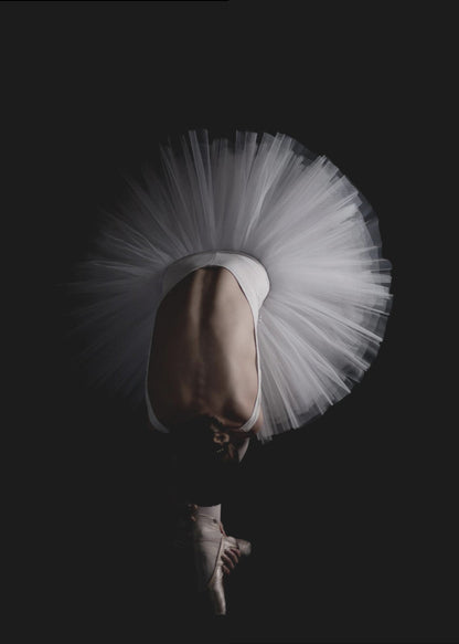 A dramatic photograph of a ballerina against a solid black background. She is bent completely forward, showing her arched back to the camera. Her white tutu fans out in a wide circle around her, and one foot in a pointe shoe is visible beneath her, balanced on its tip. Print
