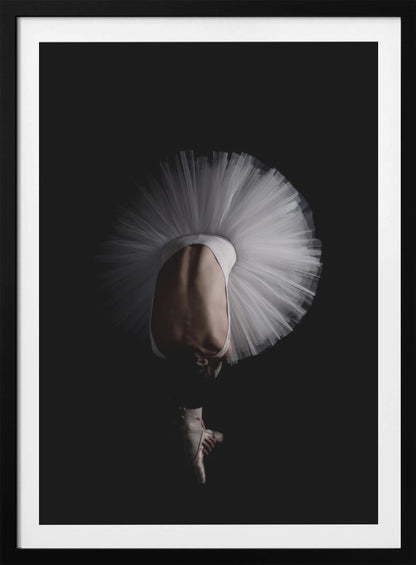 A dramatic photograph of a ballerina against a solid black background. She is bent completely forward, showing her arched back to the camera. Her white tutu fans out in a wide circle around her, and one foot in a pointe shoe is visible beneath her, balanced on its tip. Print