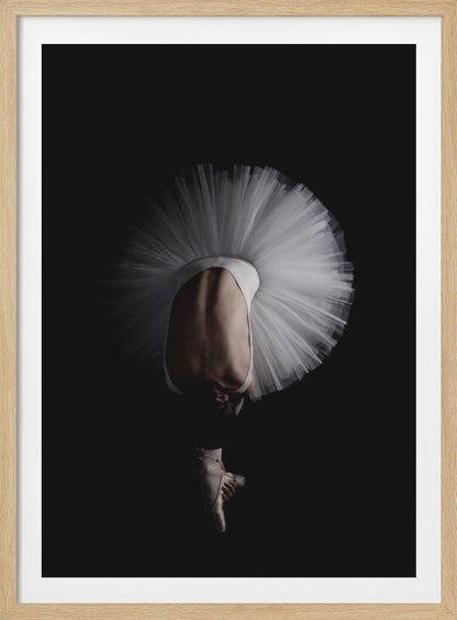 A dramatic photograph of a ballerina against a solid black background. She is bent completely forward, showing her arched back to the camera. Her white tutu fans out in a wide circle around her, and one foot in a pointe shoe is visible beneath her, balanced on its tip. Print