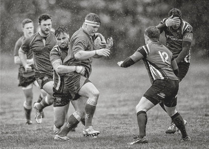 A dynamic black and white photograph of a rugby match being played in the rain. One player charges forward with the ball while an opponent wraps his arms around him in a tackle. Other players are in motion around them, capturing the intensity and physicality of the sport. Decor