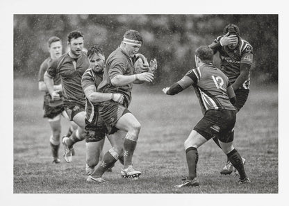 A dynamic black and white photograph of a rugby match being played in the rain. One player charges forward with the ball while an opponent wraps his arms around him in a tackle. Other players are in motion around them, capturing the intensity and physicality of the sport. Decor