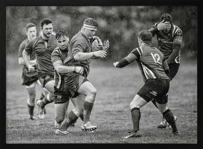 A dynamic black and white photograph of a rugby match being played in the rain. One player charges forward with the ball while an opponent wraps his arms around him in a tackle. Other players are in motion around them, capturing the intensity and physicality of the sport. Decor