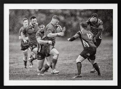 A dynamic black and white photograph of a rugby match being played in the rain. One player charges forward with the ball while an opponent wraps his arms around him in a tackle. Other players are in motion around them, capturing the intensity and physicality of the sport. Decor