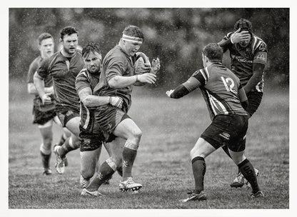 A dynamic black and white photograph of a rugby match being played in the rain. One player charges forward with the ball while an opponent wraps his arms around him in a tackle. Other players are in motion around them, capturing the intensity and physicality of the sport. Decor