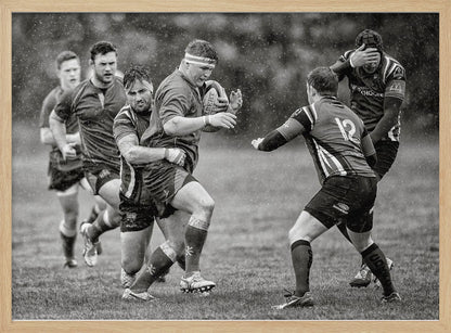 A dynamic black and white photograph of a rugby match being played in the rain. One player charges forward with the ball while an opponent wraps his arms around him in a tackle. Other players are in motion around them, capturing the intensity and physicality of the sport. Decor