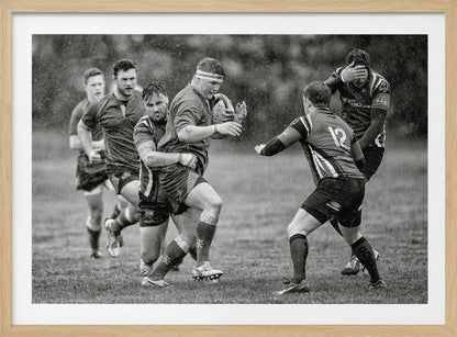 A dynamic black and white photograph of a rugby match being played in the rain. One player charges forward with the ball while an opponent wraps his arms around him in a tackle. Other players are in motion around them, capturing the intensity and physicality of the sport. Decor