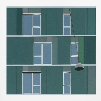 A framed photograph of a modern, dark green building facade with a grid of windows across three levels. A single industrial-style pendant lamp hangs in front of the building on the right side, interrupting the symmetrical pattern. The entire scene is enclosed by a light wood frame. Decor