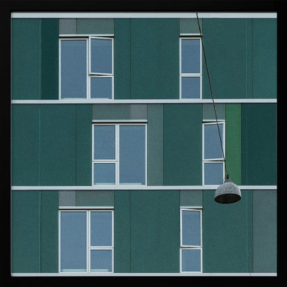 A framed photograph of a modern, dark green building facade with a grid of windows across three levels. A single industrial-style pendant lamp hangs in front of the building on the right side, interrupting the symmetrical pattern. The entire scene is enclosed by a light wood frame. Decor