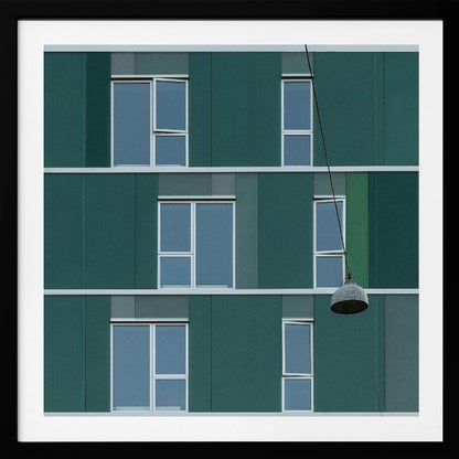 A framed photograph of a modern, dark green building facade with a grid of windows across three levels. A single industrial-style pendant lamp hangs in front of the building on the right side, interrupting the symmetrical pattern. The entire scene is enclosed by a light wood frame. Decor