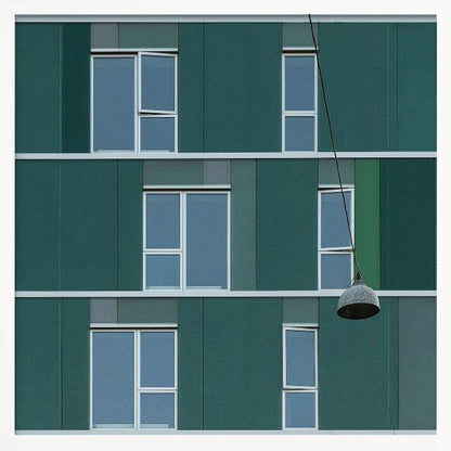 A framed photograph of a modern, dark green building facade with a grid of windows across three levels. A single industrial-style pendant lamp hangs in front of the building on the right side, interrupting the symmetrical pattern. The entire scene is enclosed by a light wood frame. Decor