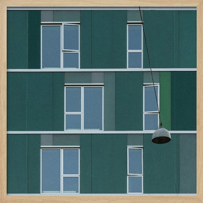 A framed photograph of a modern, dark green building facade with a grid of windows across three levels. A single industrial-style pendant lamp hangs in front of the building on the right side, interrupting the symmetrical pattern. The entire scene is enclosed by a light wood frame. Decor