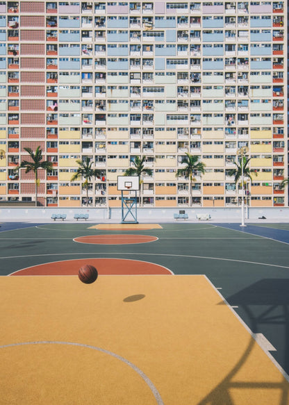 A vibrant photograph of a basketball court with a yellow and green floor, a single basketball is mid-bounce in the foreground. In the background stands the massive, symmetrical Choi Hung Estate apartment building in Hong Kong, famous for its facade of pastel rainbow colors. Palm trees line the space between the court and the building. Print