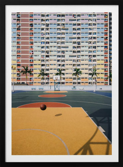 A vibrant photograph of a basketball court with a yellow and green floor, a single basketball is mid-bounce in the foreground. In the background stands the massive, symmetrical Choi Hung Estate apartment building in Hong Kong, famous for its facade of pastel rainbow colors. Palm trees line the space between the court and the building. Print