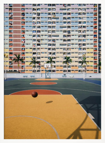 A vibrant photograph of a basketball court with a yellow and green floor, a single basketball is mid-bounce in the foreground. In the background stands the massive, symmetrical Choi Hung Estate apartment building in Hong Kong, famous for its facade of pastel rainbow colors. Palm trees line the space between the court and the building. Print