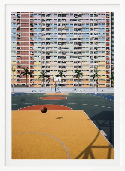 A vibrant photograph of a basketball court with a yellow and green floor, a single basketball is mid-bounce in the foreground. In the background stands the massive, symmetrical Choi Hung Estate apartment building in Hong Kong, famous for its facade of pastel rainbow colors. Palm trees line the space between the court and the building. Print