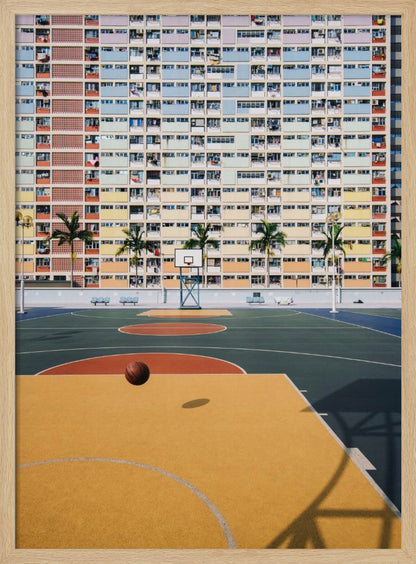 A vibrant photograph of a basketball court with a yellow and green floor, a single basketball is mid-bounce in the foreground. In the background stands the massive, symmetrical Choi Hung Estate apartment building in Hong Kong, famous for its facade of pastel rainbow colors. Palm trees line the space between the court and the building. Print