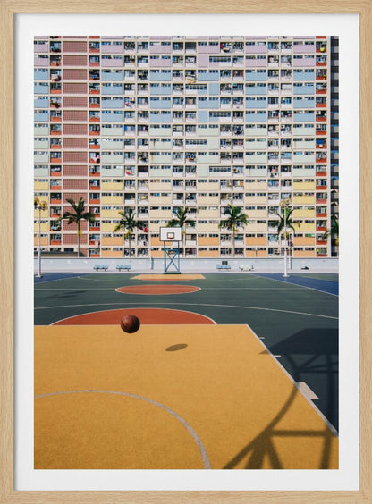 A vibrant photograph of a basketball court with a yellow and green floor, a single basketball is mid-bounce in the foreground. In the background stands the massive, symmetrical Choi Hung Estate apartment building in Hong Kong, famous for its facade of pastel rainbow colors. Palm trees line the space between the court and the building. Print