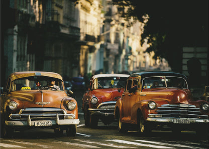 Three vintage American cars drive down a sunlit city street. The foreground features an orange taxi, with a red car and another orange vehicle behind it, all under the warm glow of late afternoon light casting long shadows. Print