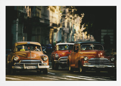 Three vintage American cars drive down a sunlit city street. The foreground features an orange taxi, with a red car and another orange vehicle behind it, all under the warm glow of late afternoon light casting long shadows. Print
