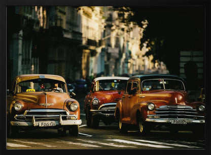 Three vintage American cars drive down a sunlit city street. The foreground features an orange taxi, with a red car and another orange vehicle behind it, all under the warm glow of late afternoon light casting long shadows. Print