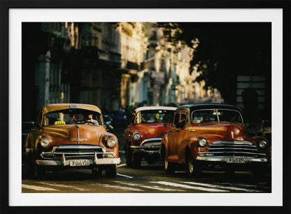 Three vintage American cars drive down a sunlit city street. The foreground features an orange taxi, with a red car and another orange vehicle behind it, all under the warm glow of late afternoon light casting long shadows. Print