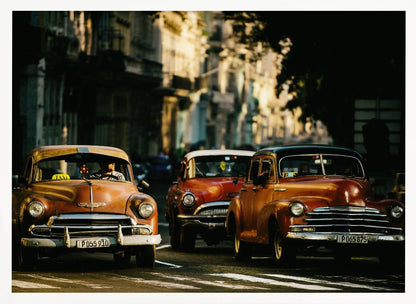 Three vintage American cars drive down a sunlit city street. The foreground features an orange taxi, with a red car and another orange vehicle behind it, all under the warm glow of late afternoon light casting long shadows. Print
