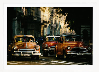 Three vintage American cars drive down a sunlit city street. The foreground features an orange taxi, with a red car and another orange vehicle behind it, all under the warm glow of late afternoon light casting long shadows. Print