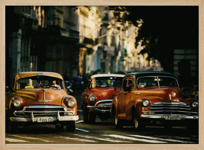 Three vintage American cars drive down a sunlit city street. The foreground features an orange taxi, with a red car and another orange vehicle behind it, all under the warm glow of late afternoon light casting long shadows. Print