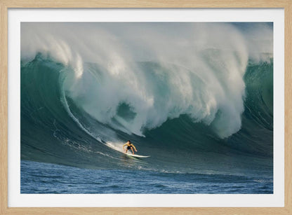 A lone surfer in a yellow shirt rides down the face of an enormous, powerful ocean wave as it crests into white foam, viewed within a silver picture frame. Wall Art