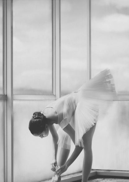 A black and white photograph of a ballerina in a white tutu, bending over to tie her pointe shoe in front of a large, bright window. The motion of her tutu is captured in a graceful blur. Print
