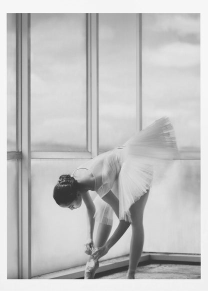 A black and white photograph of a ballerina in a white tutu, bending over to tie her pointe shoe in front of a large, bright window. The motion of her tutu is captured in a graceful blur. Print