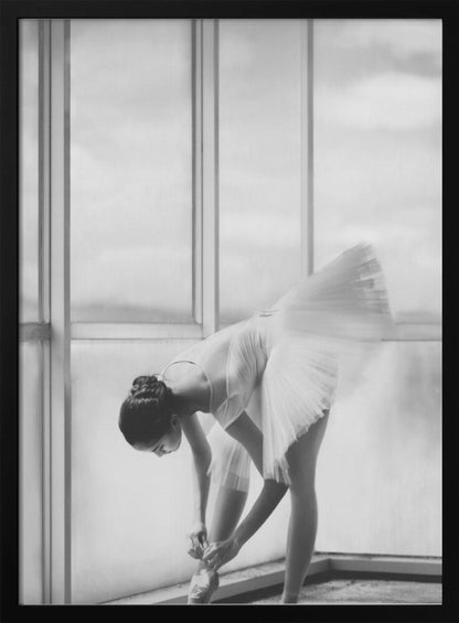 A black and white photograph of a ballerina in a white tutu, bending over to tie her pointe shoe in front of a large, bright window. The motion of her tutu is captured in a graceful blur. Print