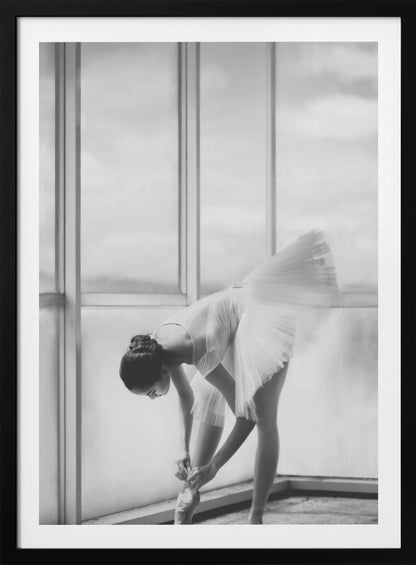A black and white photograph of a ballerina in a white tutu, bending over to tie her pointe shoe in front of a large, bright window. The motion of her tutu is captured in a graceful blur. Print