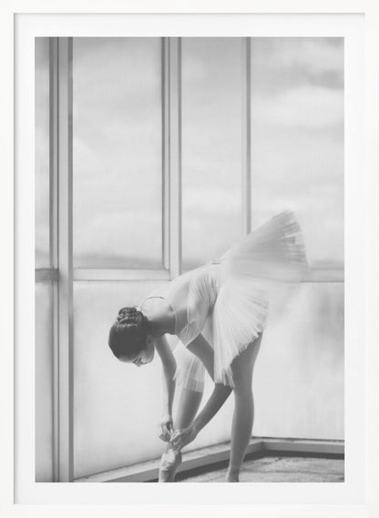 A black and white photograph of a ballerina in a white tutu, bending over to tie her pointe shoe in front of a large, bright window. The motion of her tutu is captured in a graceful blur. Print
