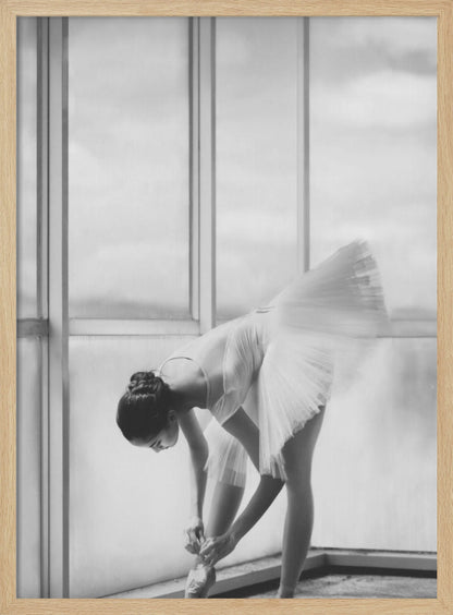 A black and white photograph of a ballerina in a white tutu, bending over to tie her pointe shoe in front of a large, bright window. The motion of her tutu is captured in a graceful blur. Print